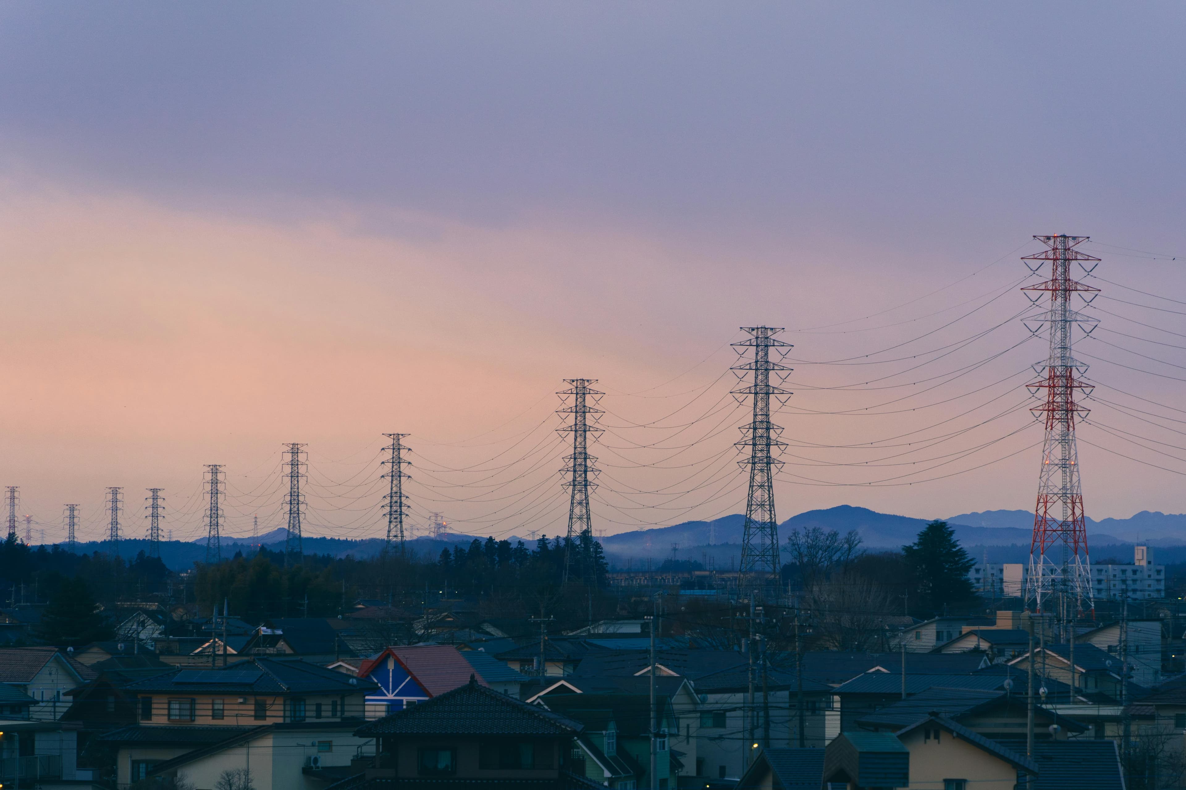 A row of high-voltage transmission towers running across a residential outer-suburb landscape at dusk, with houses in the foreground